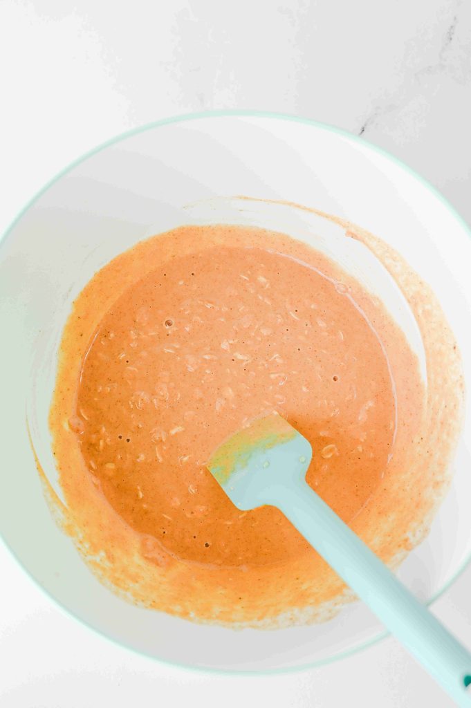 A silicone spatula stirring orange-colored high protein pumpkin overnight oats in a white mixing bowl.