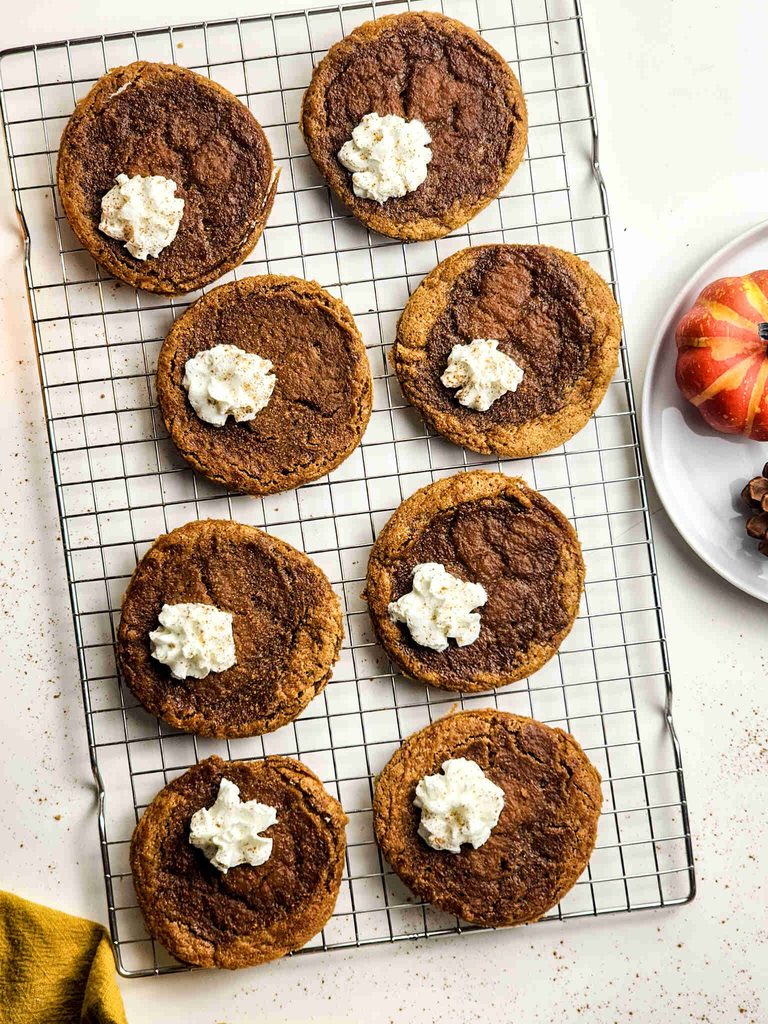 Eight pumpkin pie cookies with custard filling and whipped cream on top are arranged on a cooling rack. A pumpkin and a plate are partially visible on the right, all on a white surface.