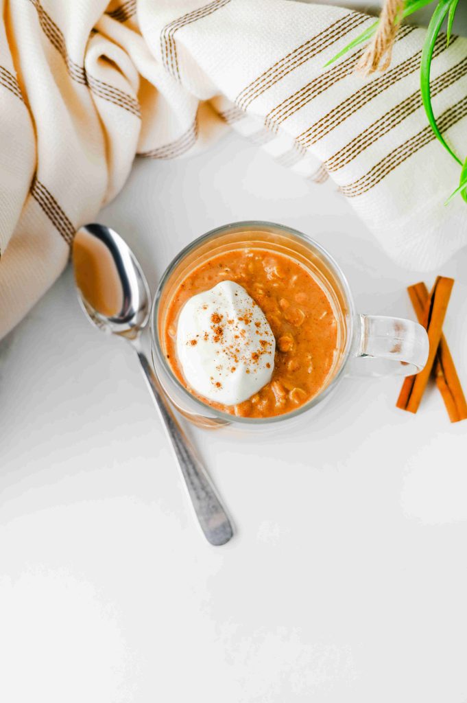 A glass mug filled with high protein pumpkin overnight oats, topped with a dollop of yogurt and a sprinkle of cinnamon, next to a spoon and cinnamon sticks.
