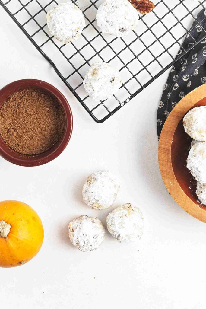 Pumpkin snowball cookies rest on a cooling rack and wooden plate, with a bowl of brown sugar and a small pumpkin arranged on a white background.