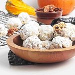 Wooden bowl filled with pumpkin snowball cookies dusted with powdered sugar, more cookies on a cooling rack behind. A small dish of pecans and a gourd are also visible.