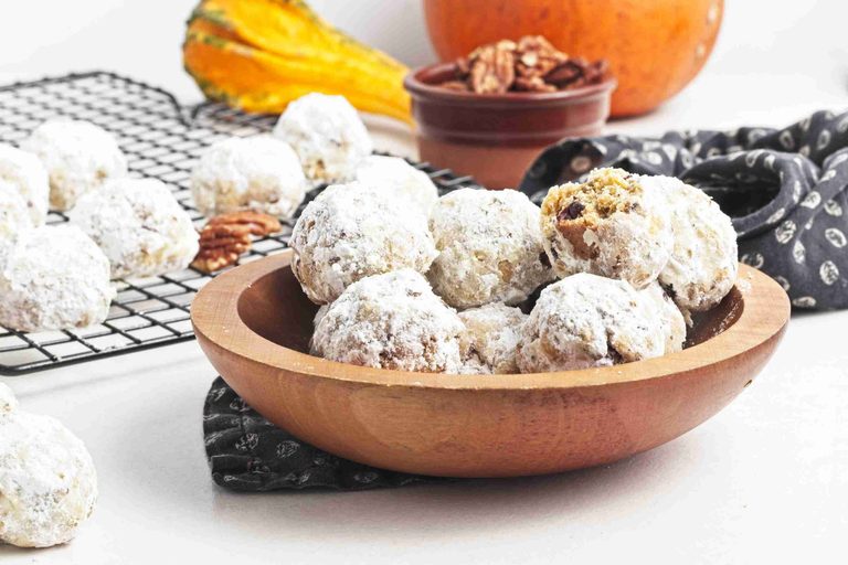 Wooden bowl filled with pumpkin snowball cookies dusted with powdered sugar, more cookies on a cooling rack behind. A small dish of pecans and a gourd are also visible.