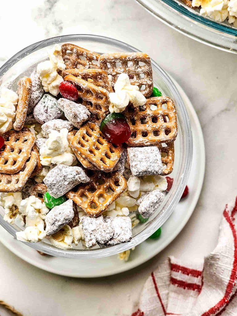 A glass bowl filled with Christmas puppy chow, a snack mix of pretzels, popcorn, powdered sugar-coated cereal, and red and green candy pieces.