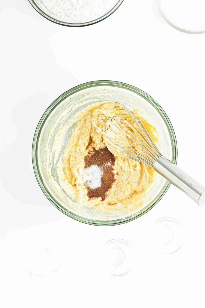 A glass bowl holds creamy batter for pumpkin snowball cookies, being mixed with a metal whisk. Cinnamon and a bit of salt rest on top, ready to be blended in. The scene is set on a white background.