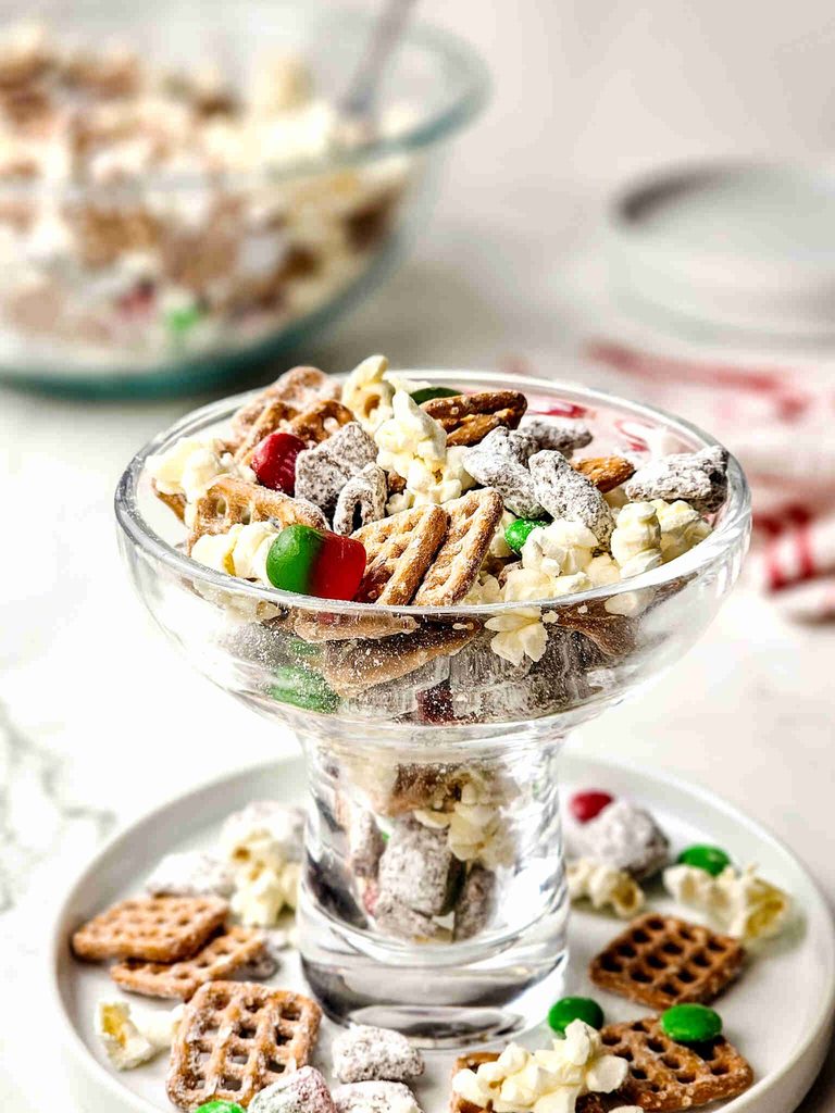 A glass pedestal bowl filled with reindeer munch, a snack mix of pretzels, popcorn, chocolate-covered cereal, and green and red candy-coated chocolates.