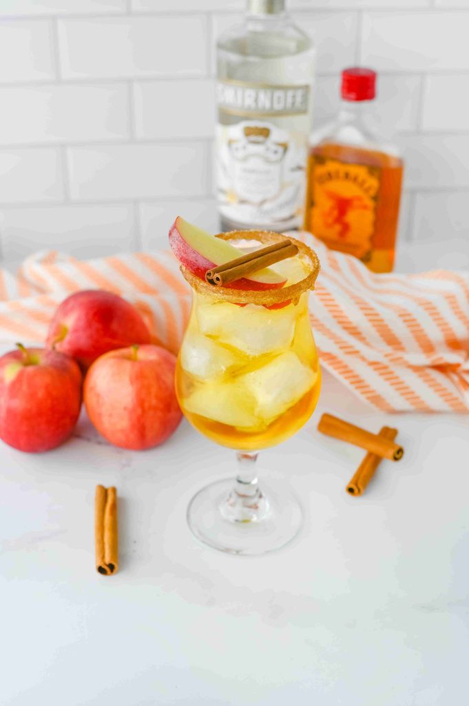 A glass of iced apple Thanksgiving vodka cocktail garnished with an apple slice and cinnamon stick, with whole apples and liquor bottles in the background.