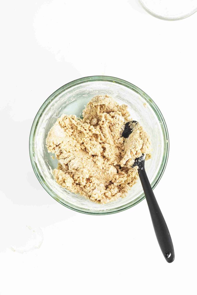 A glass bowl filled with pumpkin snowball cookie dough sits on a white surface. A black spoon is partially buried in the dough, showing it has been mixed.