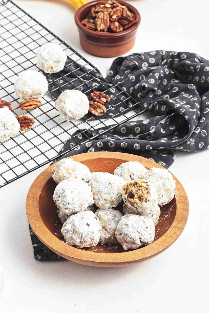 A wooden bowl of pumpkin snowball cookies sits on a dark napkin, with more cookies on a cooling rack and a small bowl of pecans nearby.